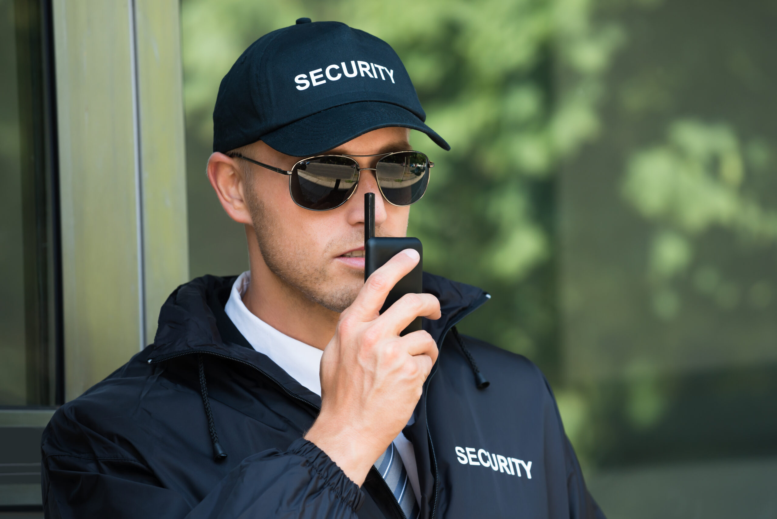A security guard in a black jacket and cap holds a walkie-talkie close to his mouth