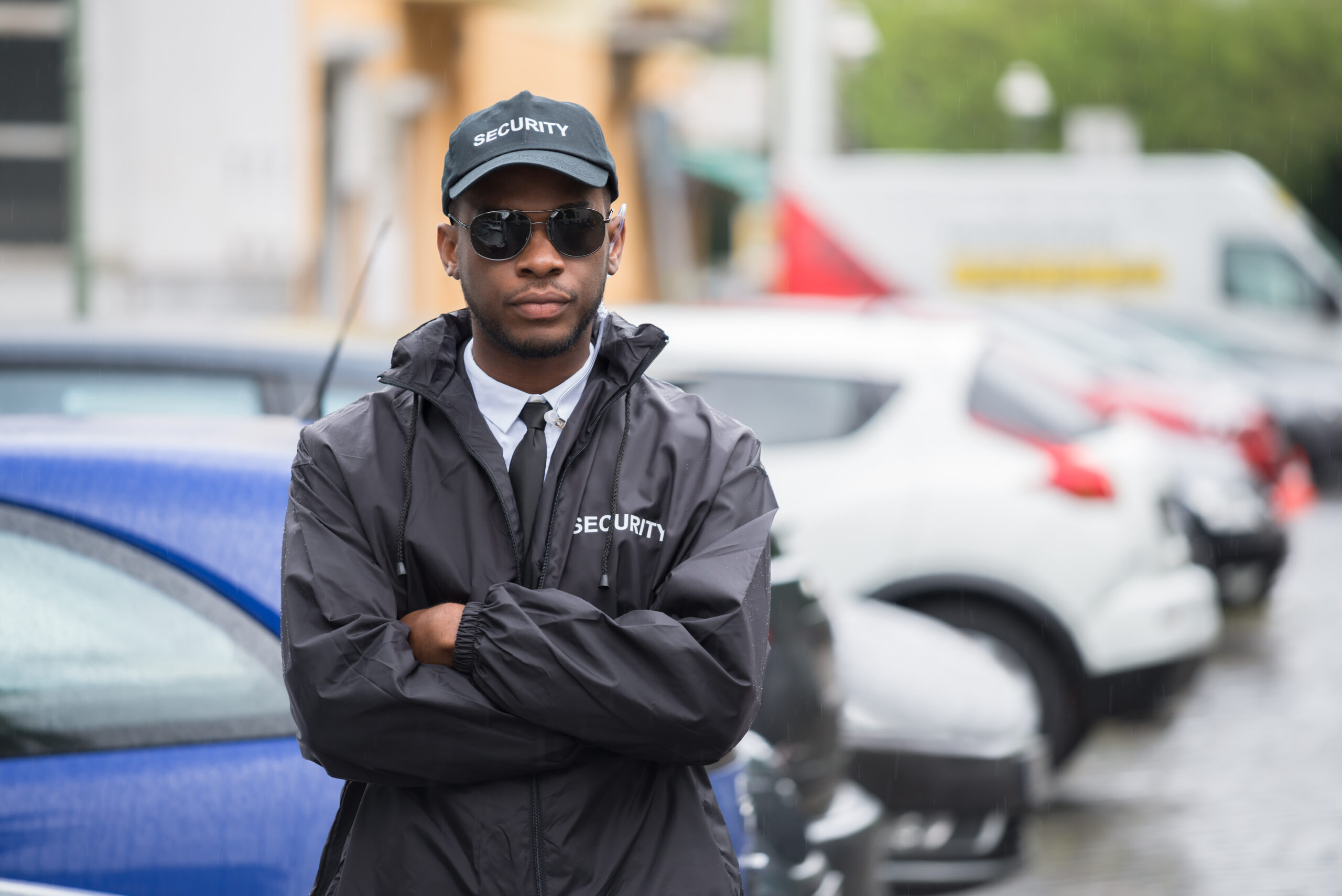 A security guard in a black jacket and cap stands with arms crossed in a parking lot